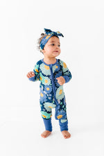 A toddler wears the Jammie Session Bluepsie Daisy Romper— a blue floral bamboo long-sleeve romper with convertible footies and matching headband— while standing against a plain white background.