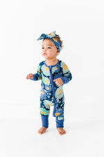 A toddler stands on a white floor wearing the Jammie Session Bluepsie Daisy Romper with convertible footies and a matching headband, looking slightly to the side.
