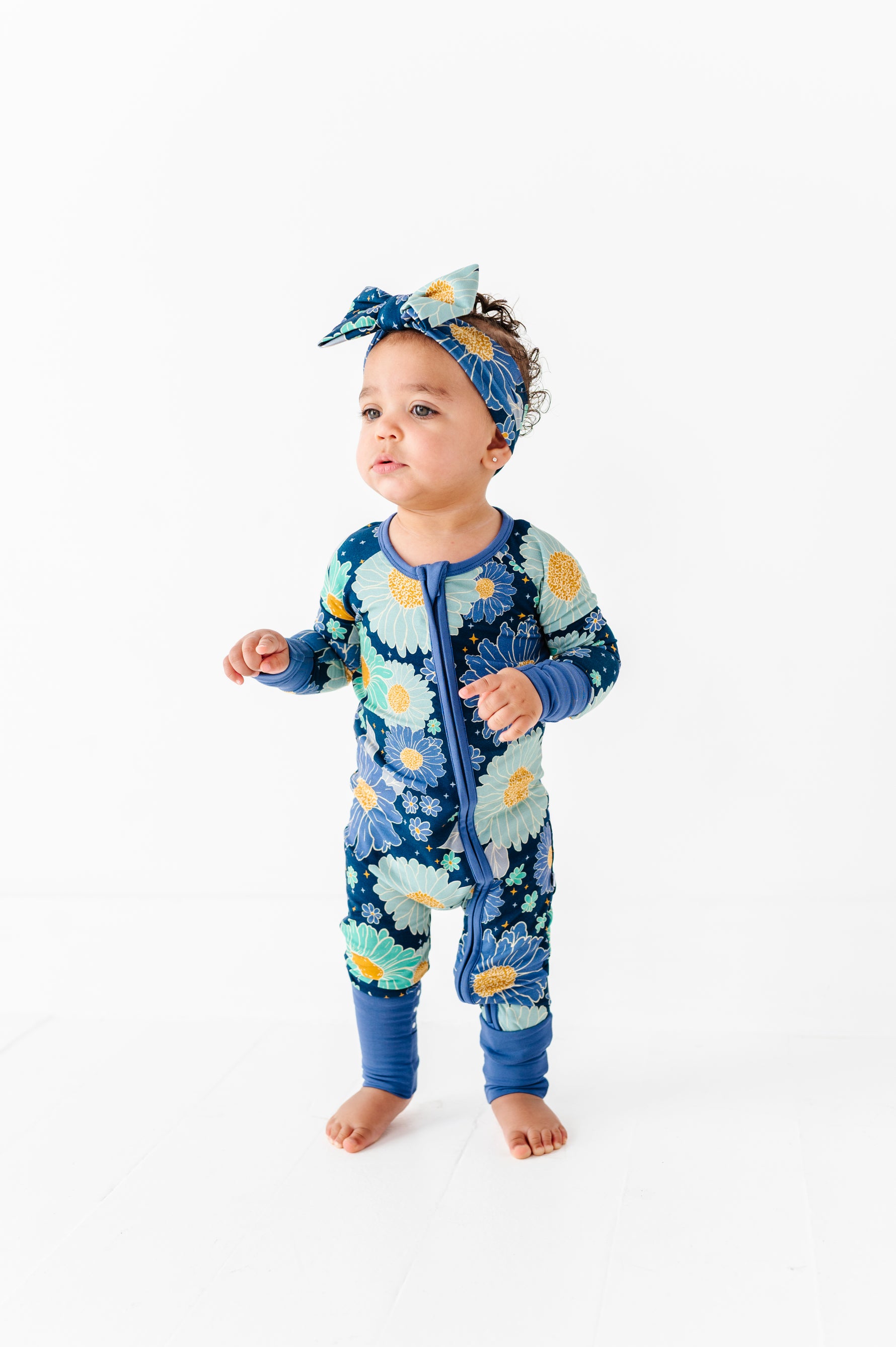A toddler stands on a white floor wearing the Jammie Session Bluepsie Daisy Romper with convertible footies and a matching headband, looking slightly to the side.