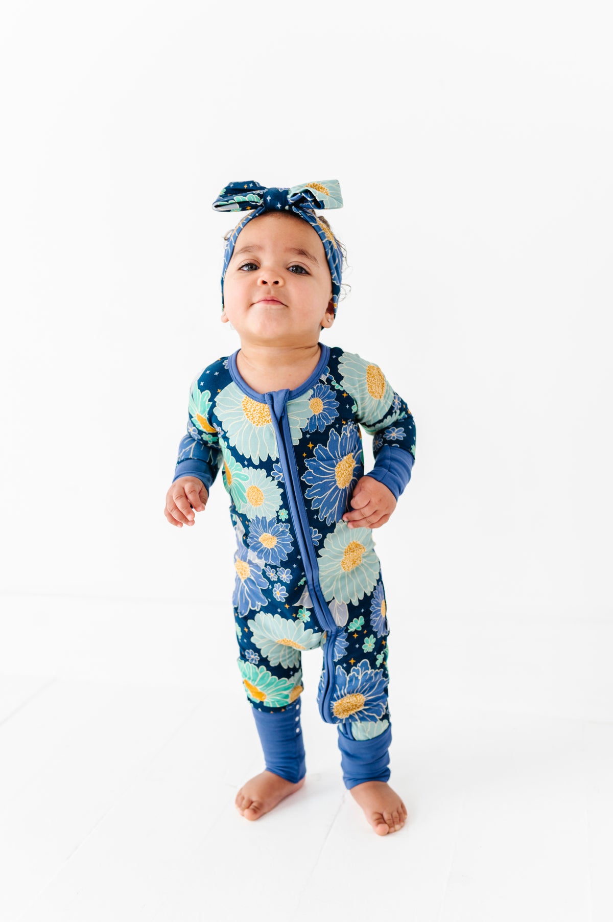 A baby wears the Jammie Session Bluepsie Daisy Romper with long sleeves, convertible footies, a matching headband, and a double zipper for easy changes, standing barefoot on a white floor against a plain white background.