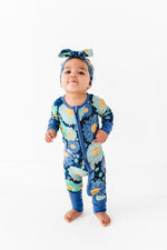 A baby wears the Jammie Session Bluepsie Daisy Romper with long sleeves, convertible footies, a matching headband, and a double zipper for easy changes, standing barefoot on a white floor against a plain white background.