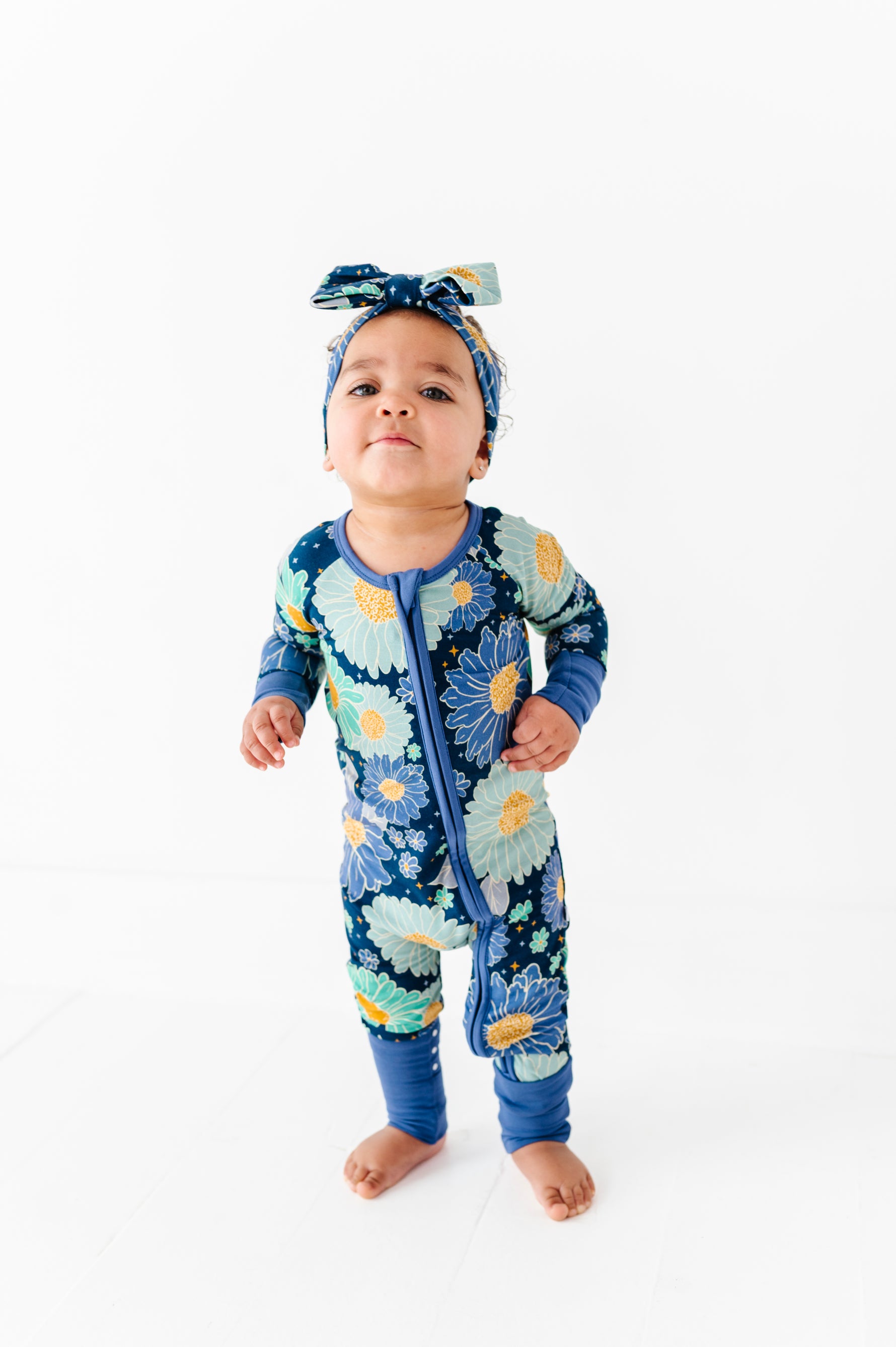 A baby wears the Jammie Session Bluepsie Daisy Romper with long sleeves, convertible footies, a matching headband, and a double zipper for easy changes, standing barefoot on a white floor against a plain white background.