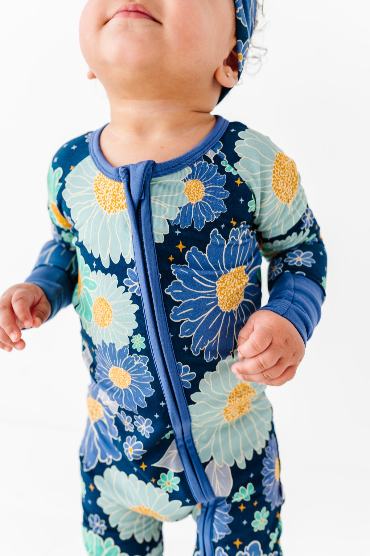A baby wears the Jammie Session Bluepsie Daisy Romper, a blue floral long-sleeve bamboo romper with convertible footies and matching headband, standing against a white background.