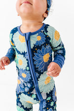 A baby wears the Jammie Session Bluepsie Daisy Romper, a blue floral long-sleeve bamboo romper with convertible footies and matching headband, standing against a white background.
