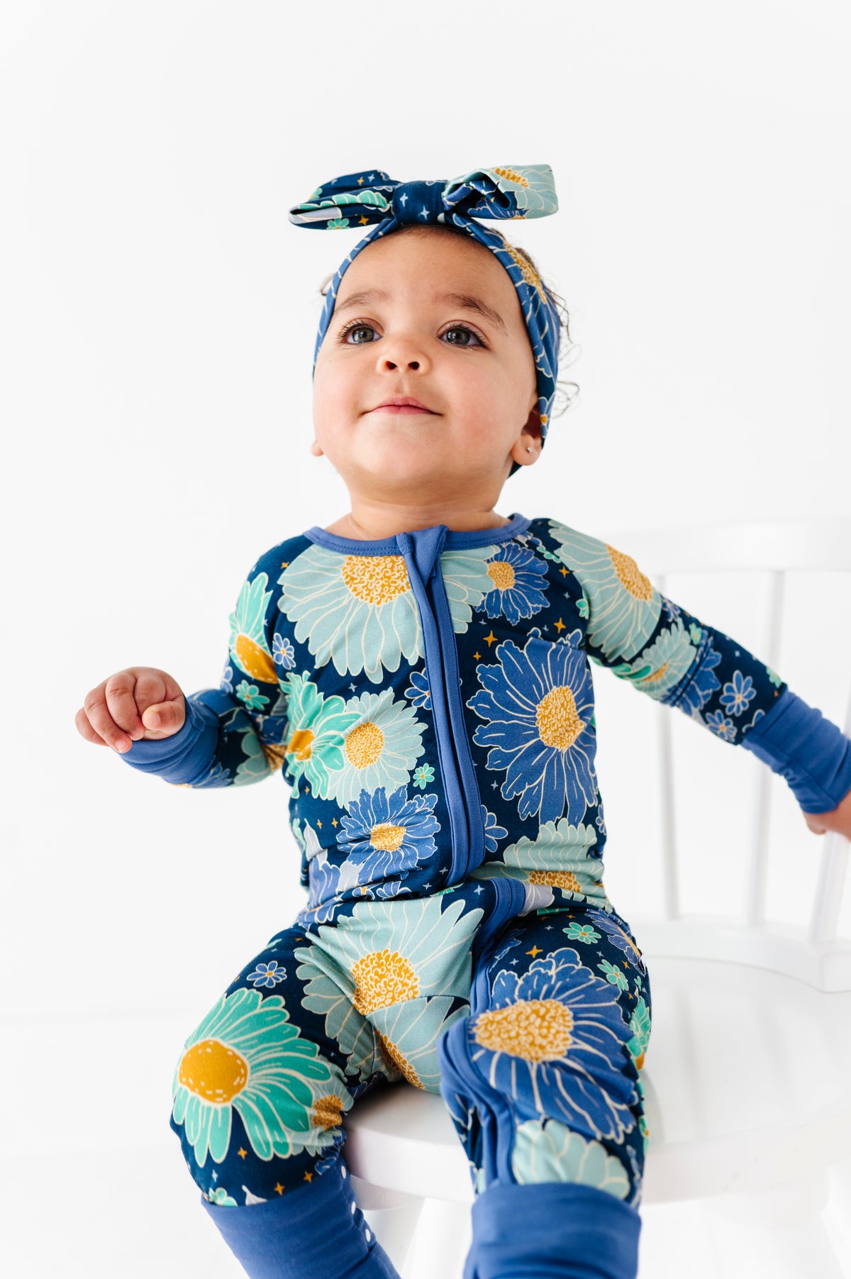 A toddler wearing the Jammie Session Bluepsie Daisy Romper with convertible footies and a matching headband sits on a white chair against a plain white background.