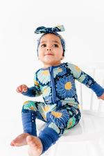 A young child wears the Jammie Session Bluepsie Daisy Romper, a blue floral long-sleeve bamboo outfit with convertible footies and a matching headband, sitting barefoot on a white chair against a white background.