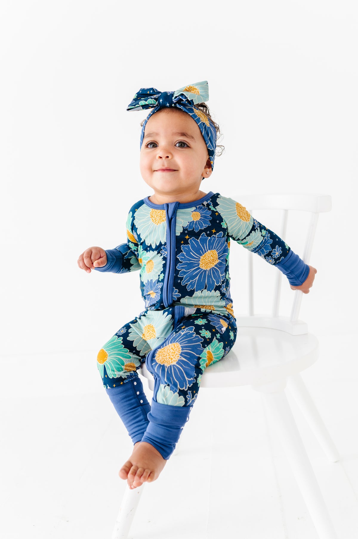 A toddler sits on a white chair, smiling and looking up, wearing the Jammie Session Bluepsie Daisy Romper with blue floral print, long sleeves, convertible footies, and a matching headband.