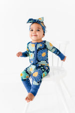 A toddler sits on a white chair, smiling and looking up, wearing the Jammie Session Bluepsie Daisy Romper with blue floral print, long sleeves, convertible footies, and a matching headband.