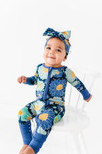 A baby in a Jammie Session Bluepsie Daisy Romper with convertible footies and matching headband sits on a white chair against a white background.