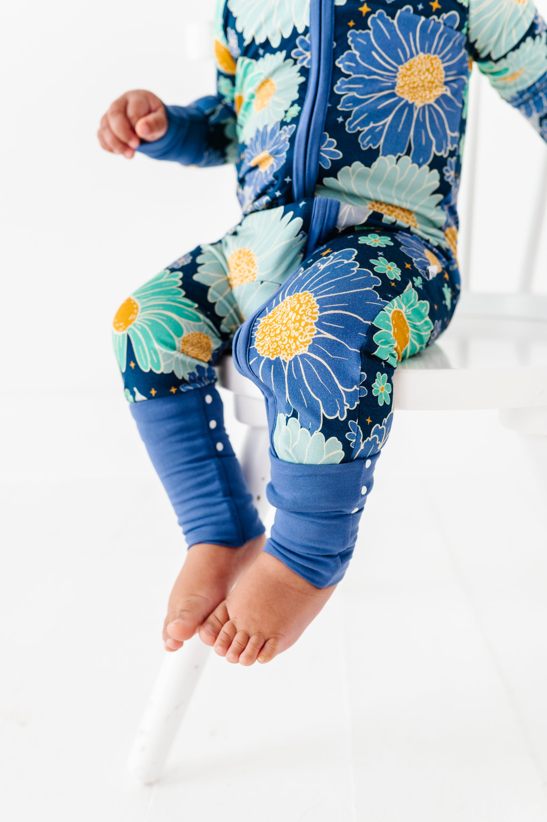 A child sits on a white chair, wearing the Jammie Session Bluepsie Daisy Romper with blue floral print, blue cuffs, and convertible footies. Only the lower half of their body is visible.