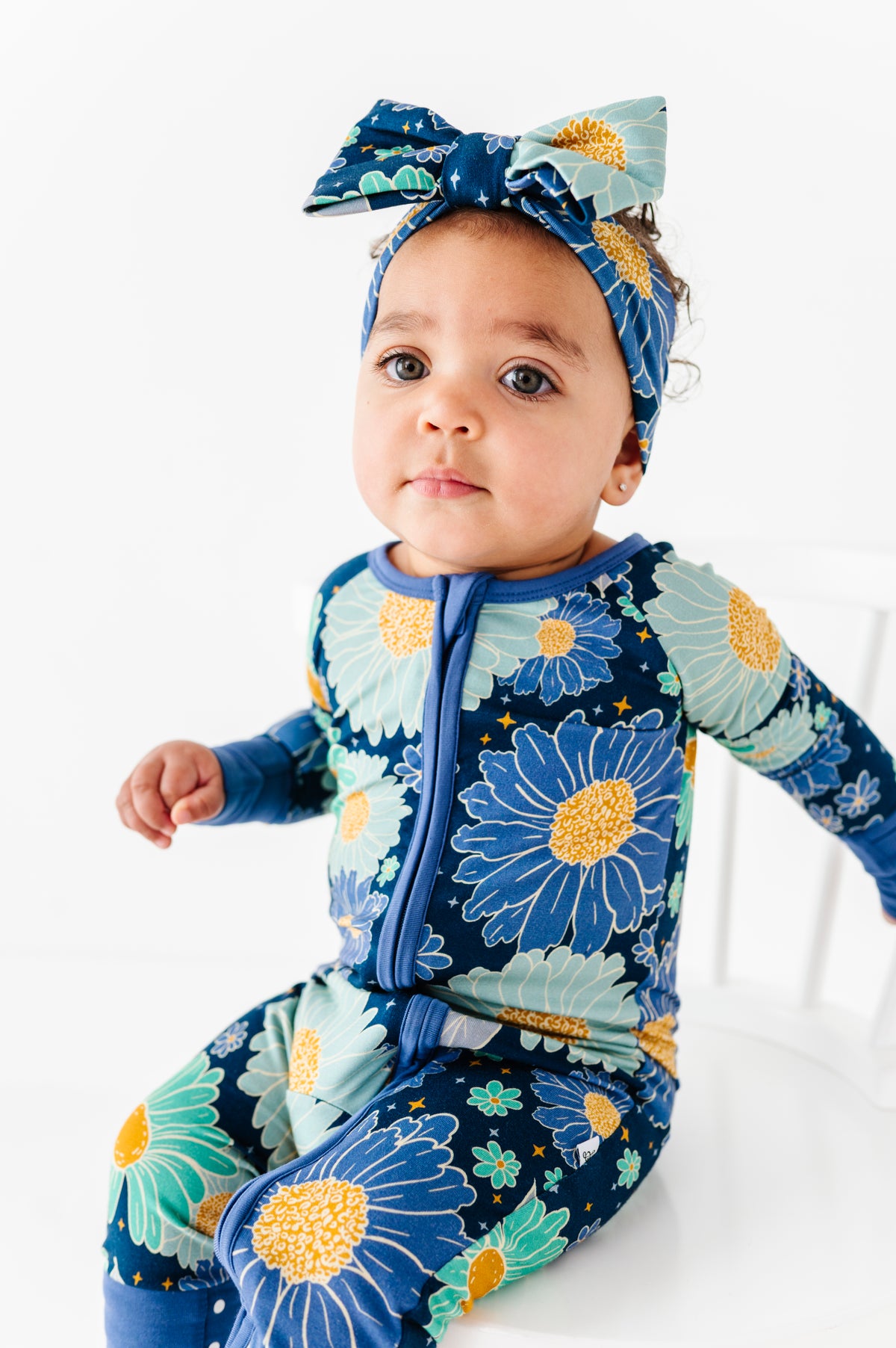 A baby wearing the Jammie Session Bluepsie Daisy Romper with blue floral pattern, convertible footies, and double zipper sits on a white chair against a plain white background.
