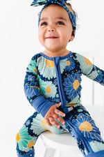 A baby in the Jammie Session Bluepsie Daisy Romper with convertible footies and a matching headband sits on a white chair, smiling and looking slightly upward.