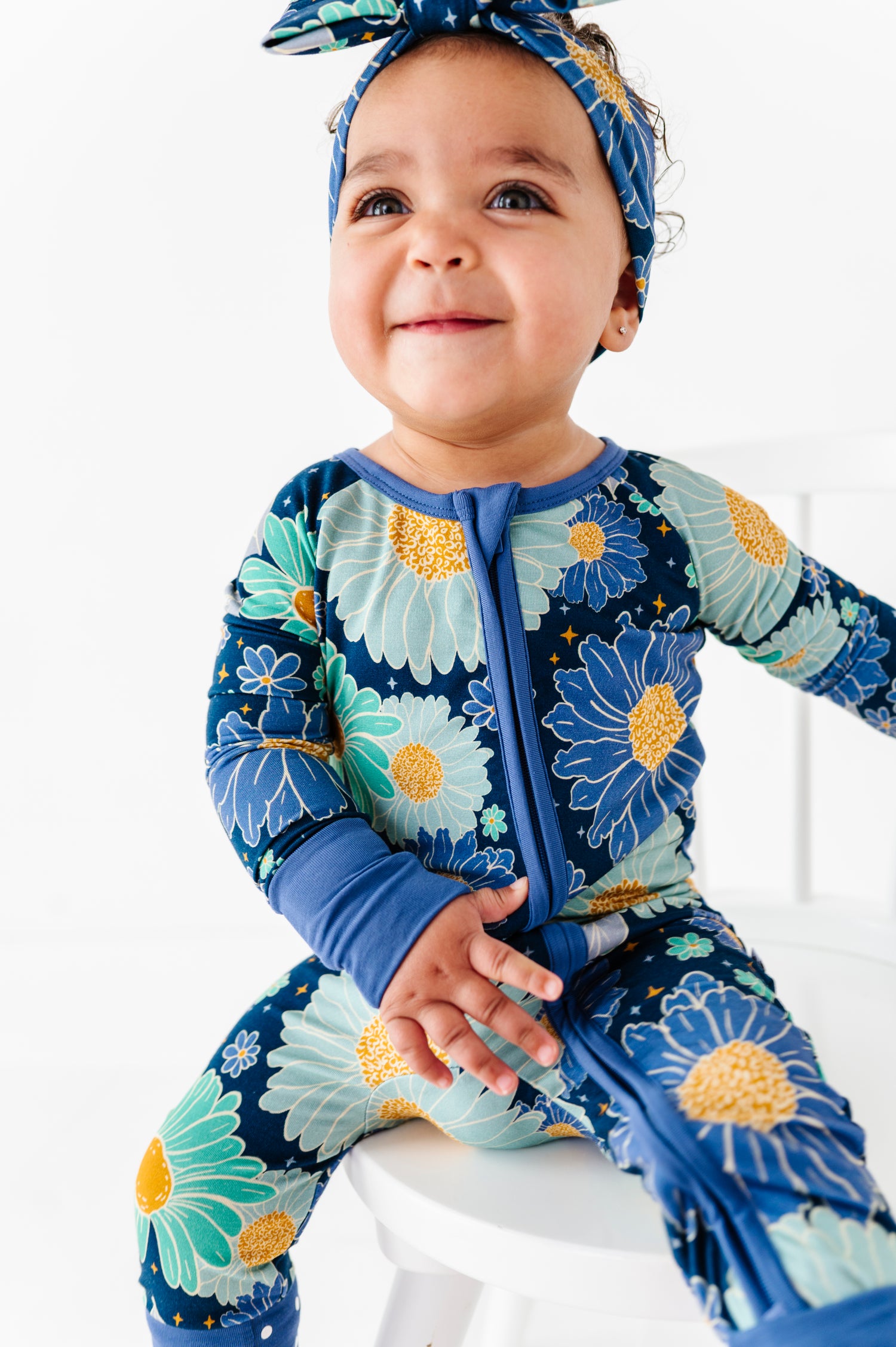 A baby in the Jammie Session Bluepsie Daisy Romper with convertible footies and a matching headband sits on a white chair, smiling and looking slightly upward.