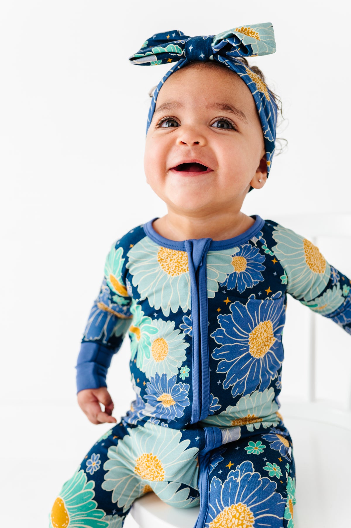 A baby sits smiling against a white background, wearing the Jammie Session Bluepsie Daisy Romper—a blue floral-patterned bamboo long sleeve romper with convertible footies—and a matching headband.