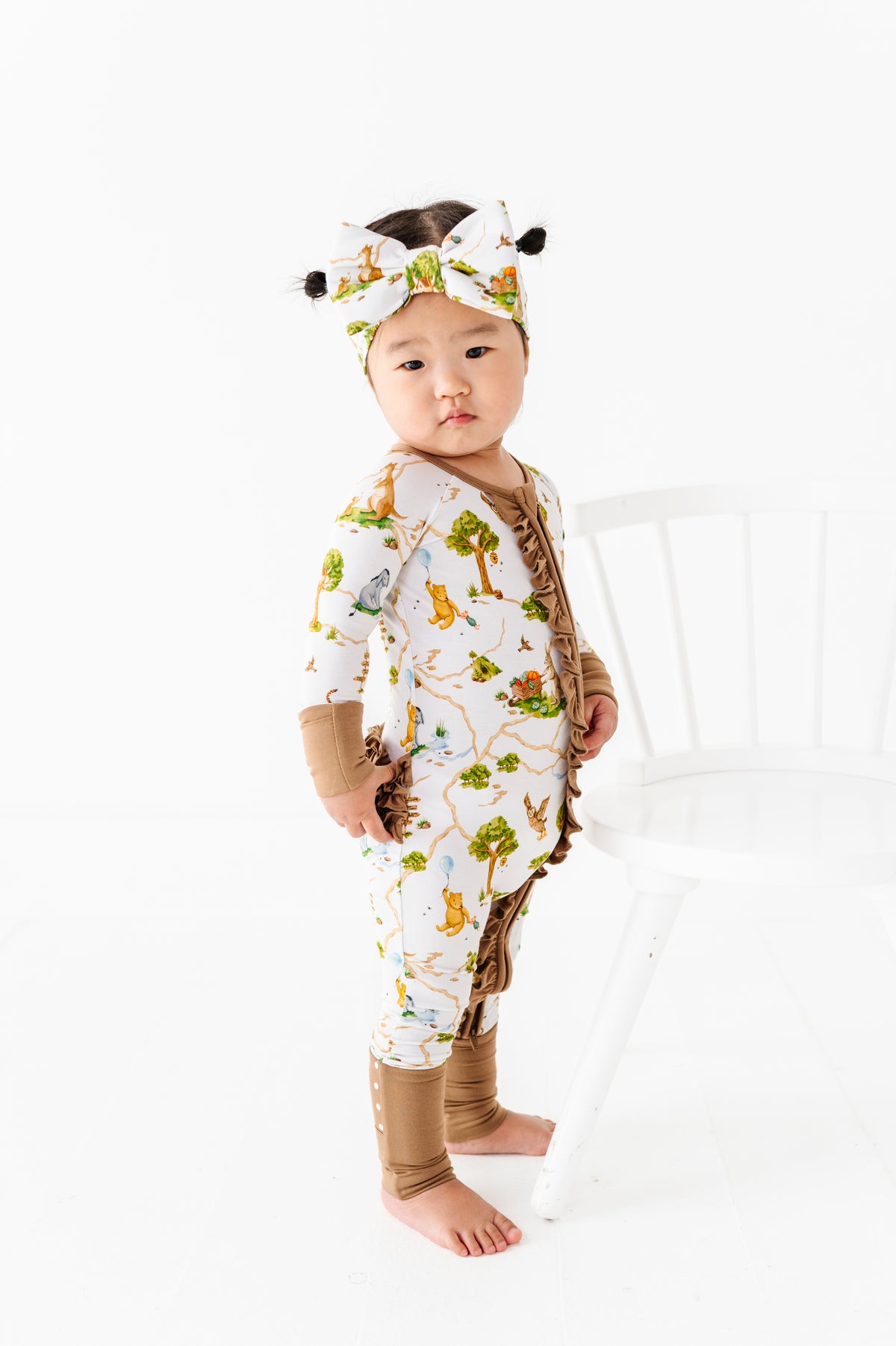 A baby in the Oh Bother, It's Bedtime Ruffle Romper by Jammie Session, featuring an animal print and matching headband, stands beside a white chair on a white background.