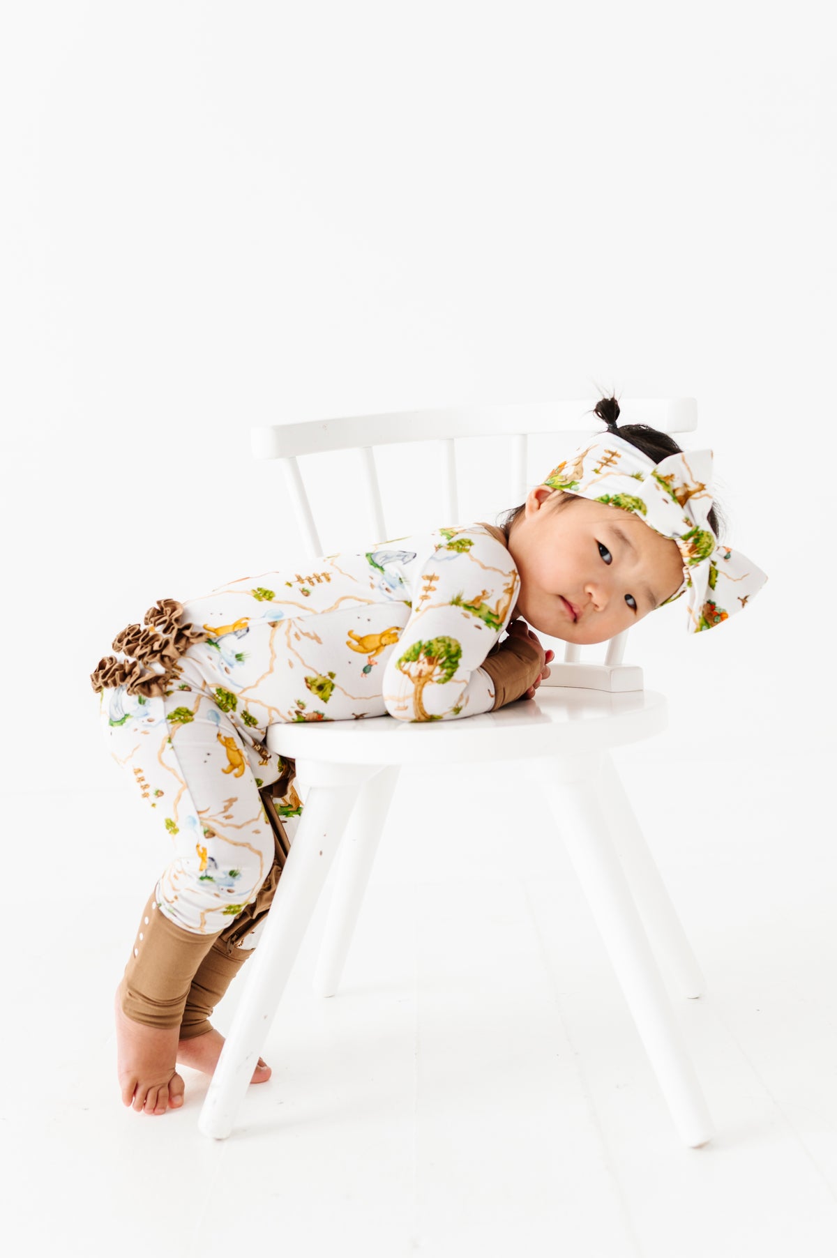 A toddler wears the Jammie Session Oh Bother, It’s Bedtime Ruffle Romper with a matching headband, leaning on a white chair and looking at the camera in a bright, minimalist space.