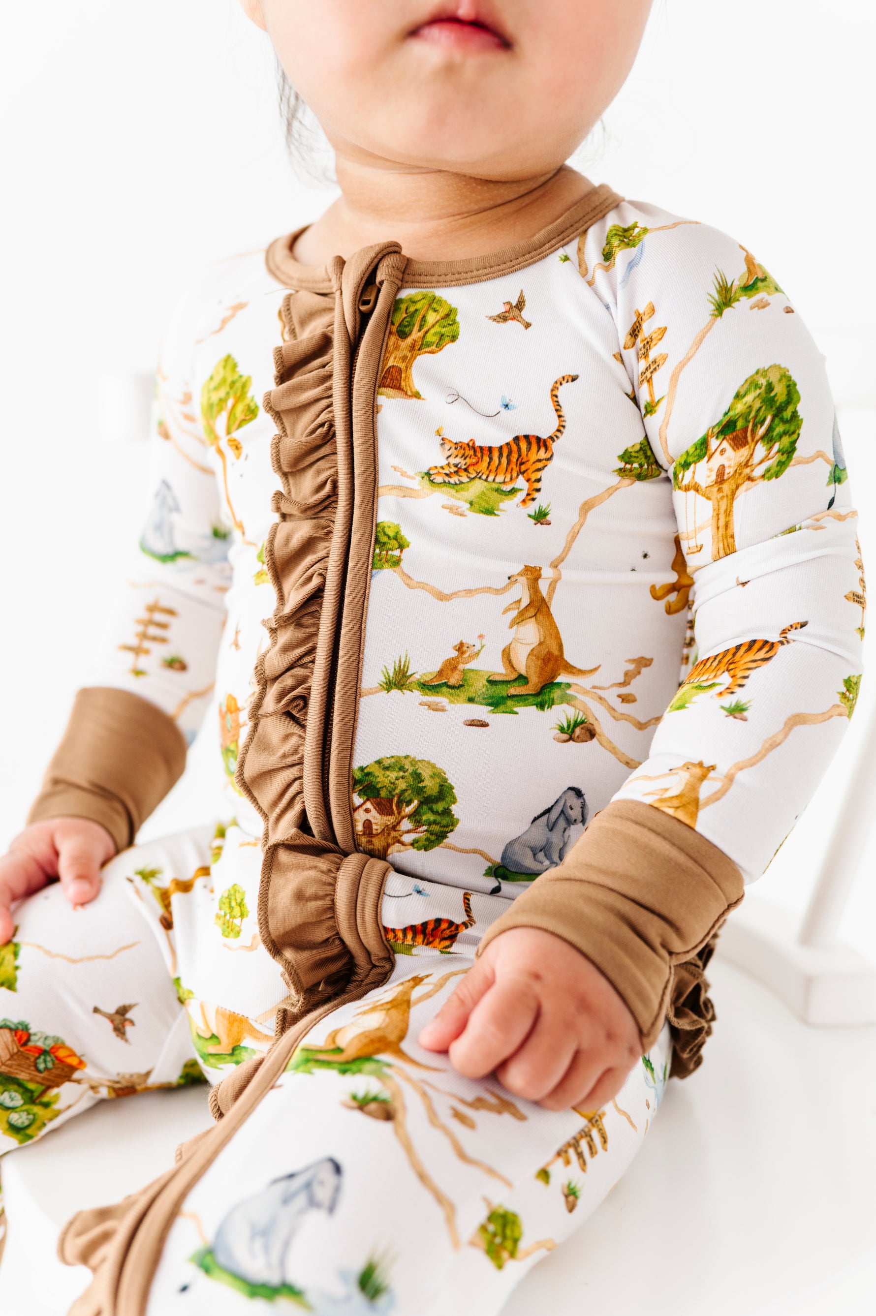 A baby in the Jammie Session "Oh Bother, It's Bedtime Ruffle Romper"—a white bamboo long sleeve romper with animal and tree prints, brown ruffle trim, and cuffs—sits against a white background.