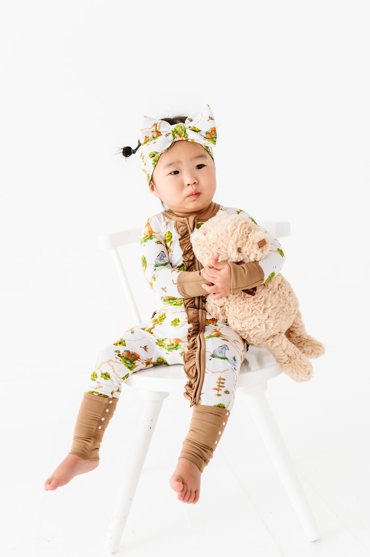 A baby sits on a white chair, wearing the Jammie Session "Oh Bother, It's Bedtime Ruffle Romper" and a headband, holding a beige stuffed animal.
