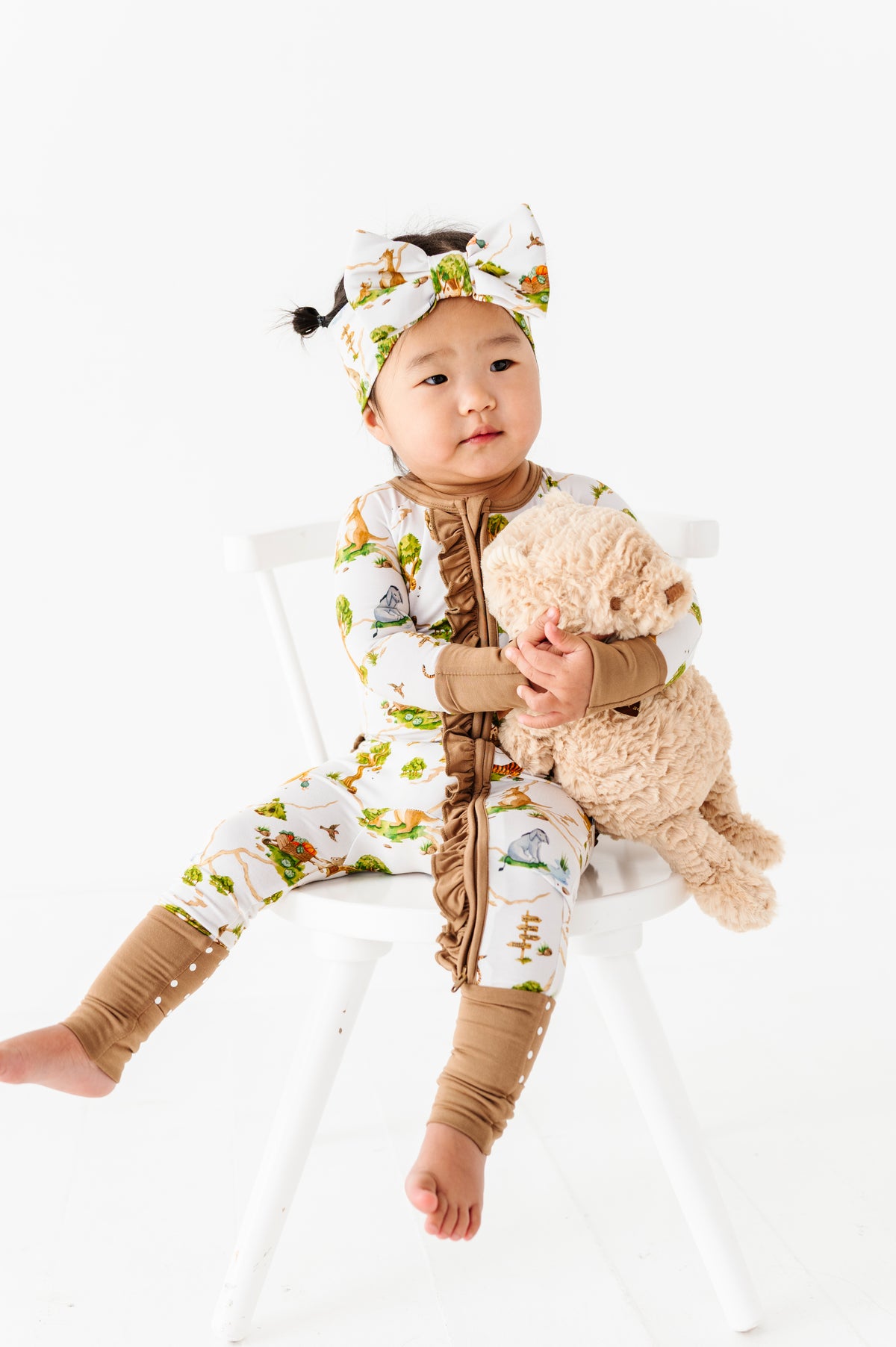 A baby wears the Jammie Session "Oh Bother, It's Bedtime" Ruffle Romper and headband while sitting on a white chair, holding a plush dog toy.