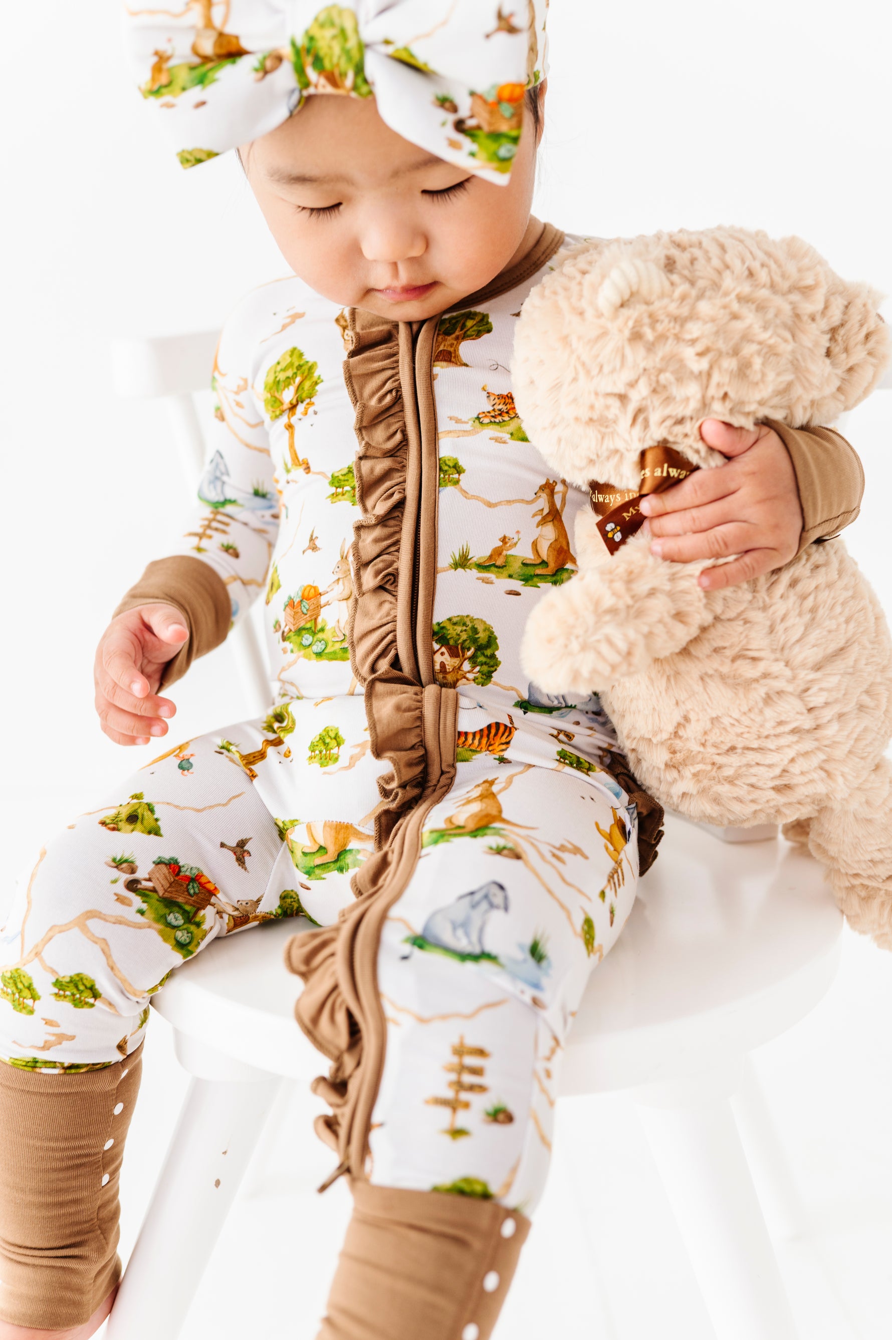 A baby wearing the Jammie Session Oh Bother, It's Bedtime Ruffle Romper and matching headband sits on a white chair, holding a plush teddy bear.
