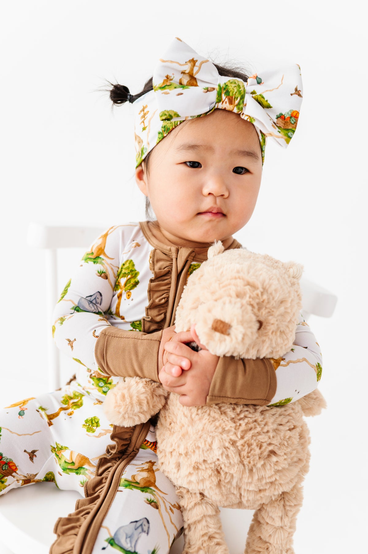 A toddler in the Jammie Session "Oh Bother, It's Bedtime" Ruffle Romper with matching headband sits on a white chair, holding a beige stuffed animal.