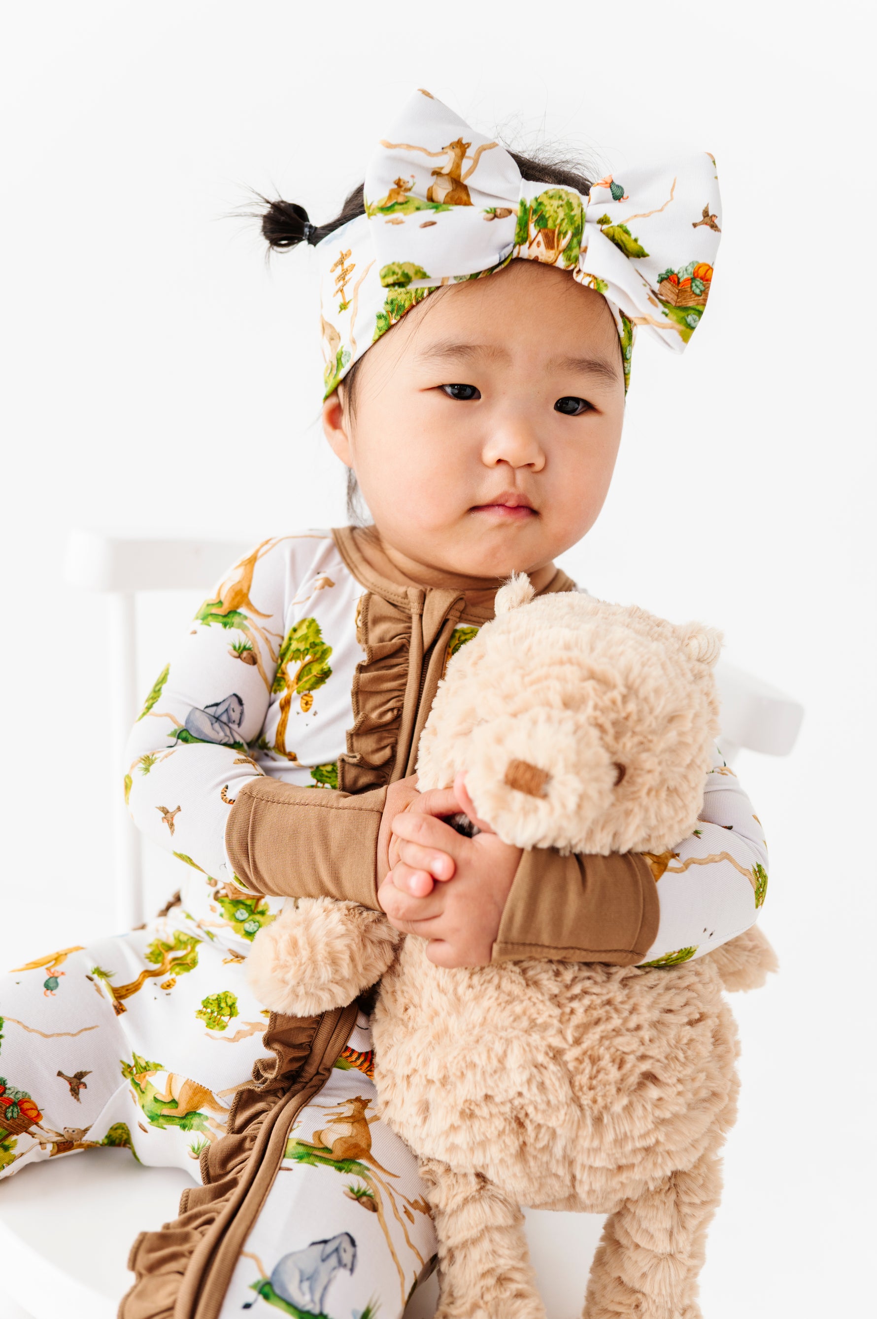 A toddler in the Jammie Session "Oh Bother, It's Bedtime" Ruffle Romper with matching headband sits on a white chair, holding a beige stuffed animal.