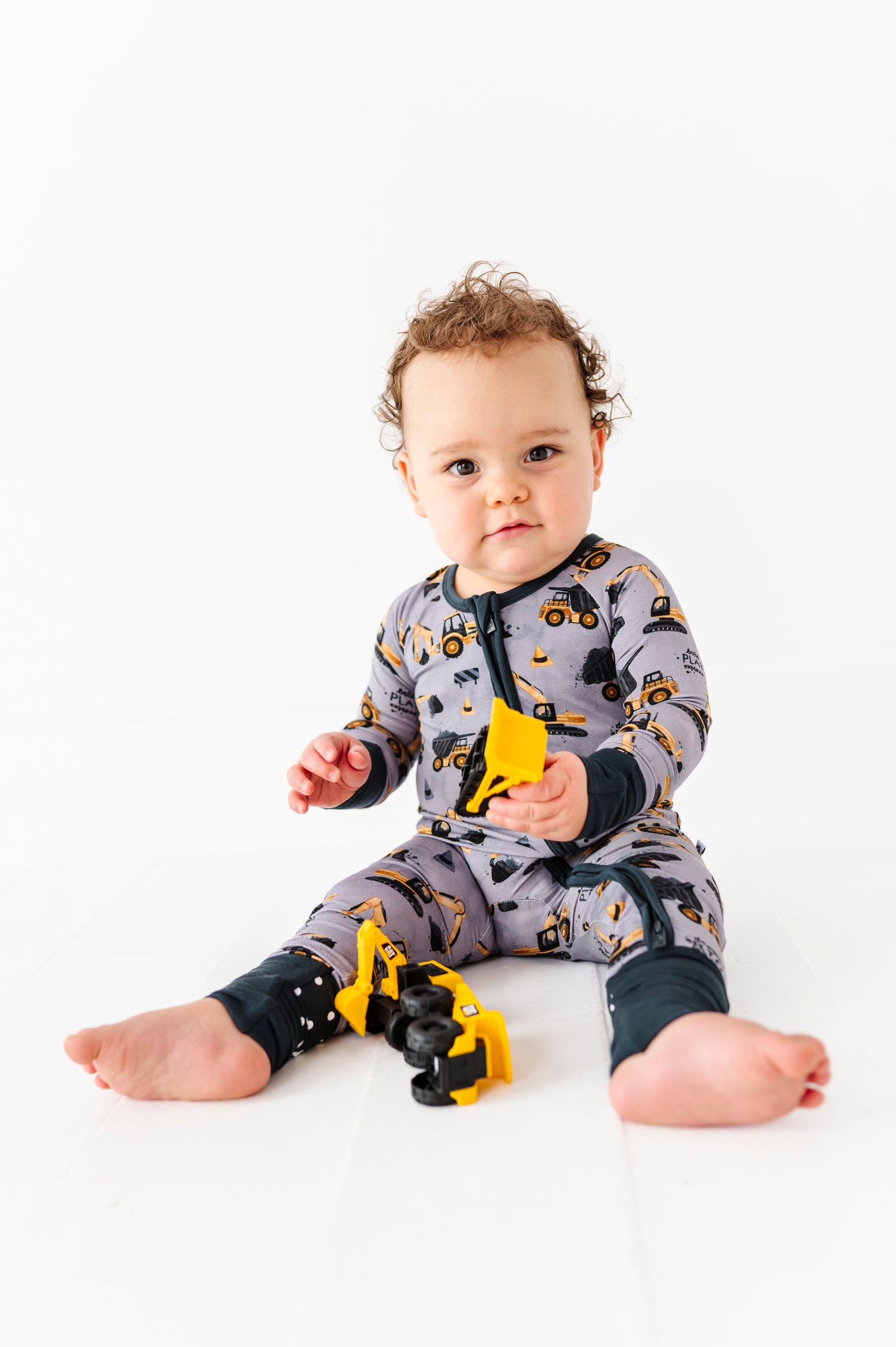 A baby wearing the Jammie Session Snooze Crew Romper with a construction vehicle theme sits on the floor, holding a yellow toy construction vehicle part.