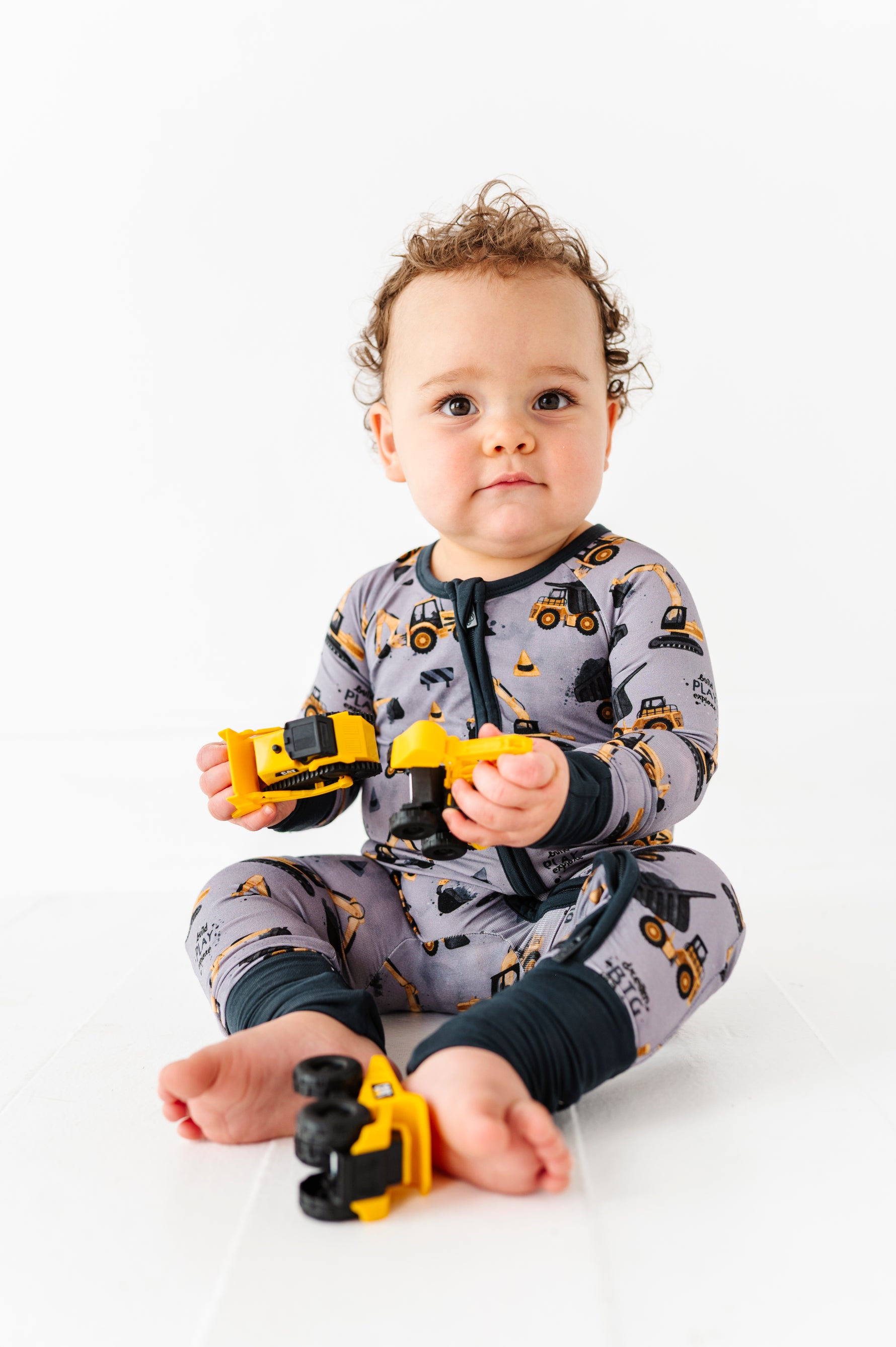 A baby wearing the Jammie Session Snooze Crew Romper featuring a construction vehicle pattern sits on the floor holding yellow toy construction trucks against a white background.