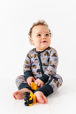 A baby in the Jammie Session Snooze Crew Romper featuring construction vehicles sits on the floor, holding a yellow toy truck and smiling.