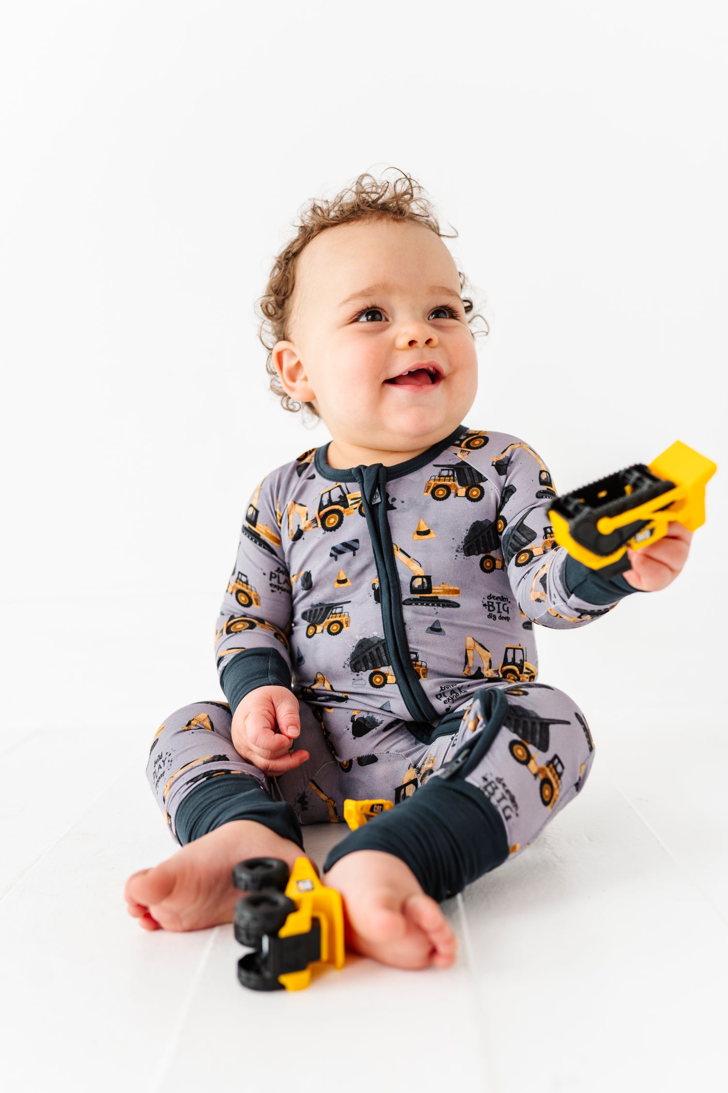 A baby in a Jammie Session Snooze Crew Romper sits on the floor, smiling and holding a yellow toy truck, with another toy truck by their feet.