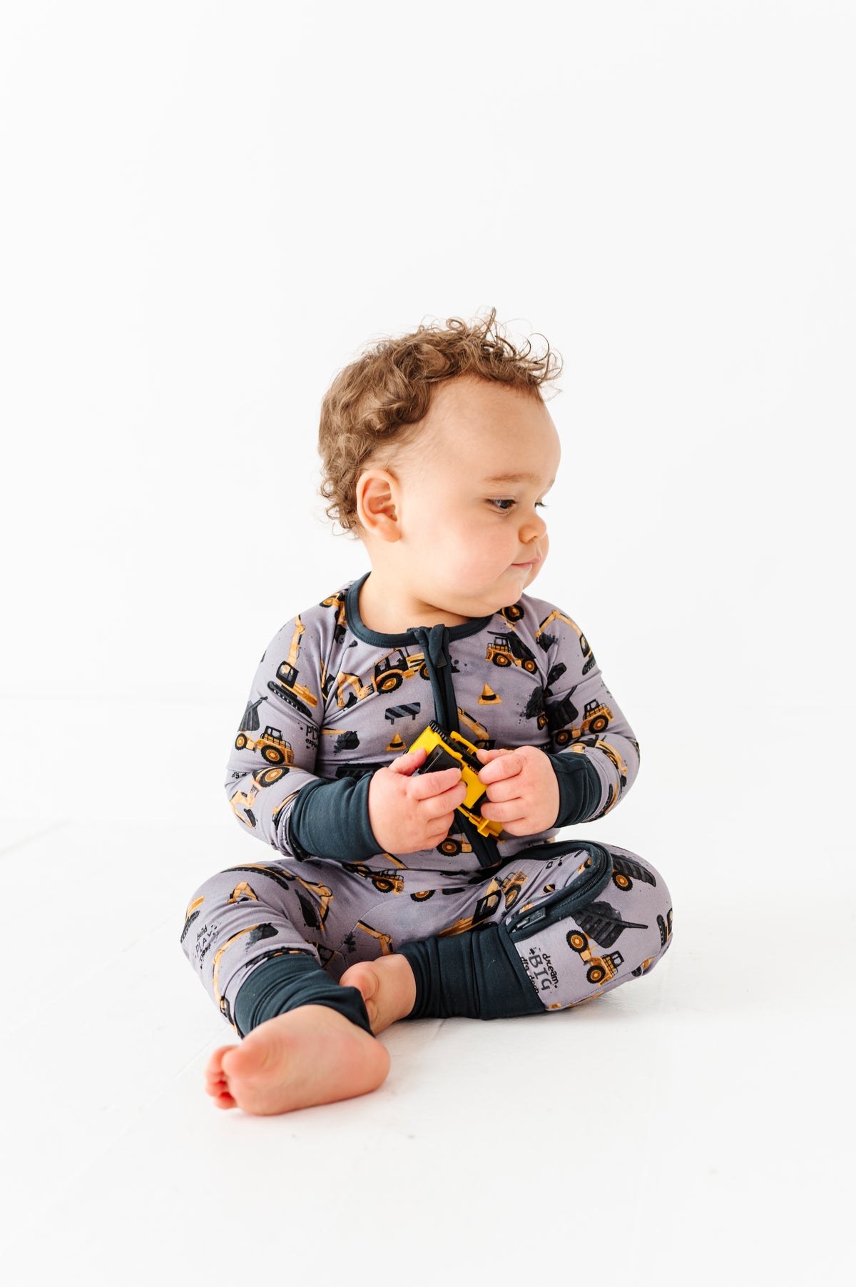 A baby sits on the floor wearing the Jammie Session Snooze Crew Romper with a construction-vehicle print, holding a small yellow toy and looking to the side against a white background.