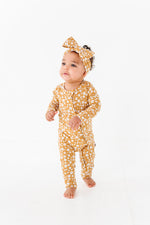 A baby in the Jammie Session Honey Blooms Romper with a matching headband stands on a white floor against a white background.