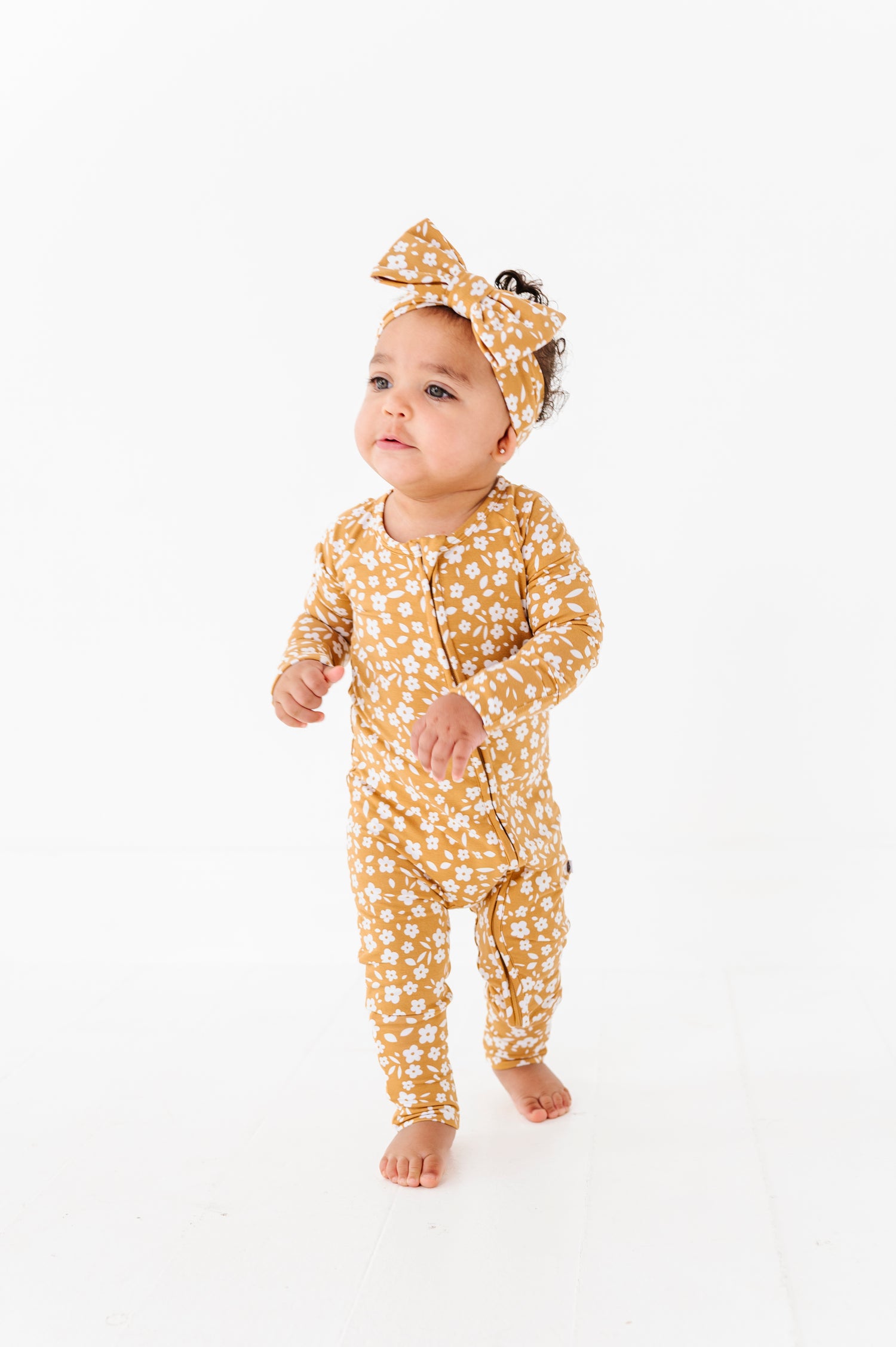 A baby in the Jammie Session Honey Blooms Romper with a matching headband stands on a white floor against a white background.