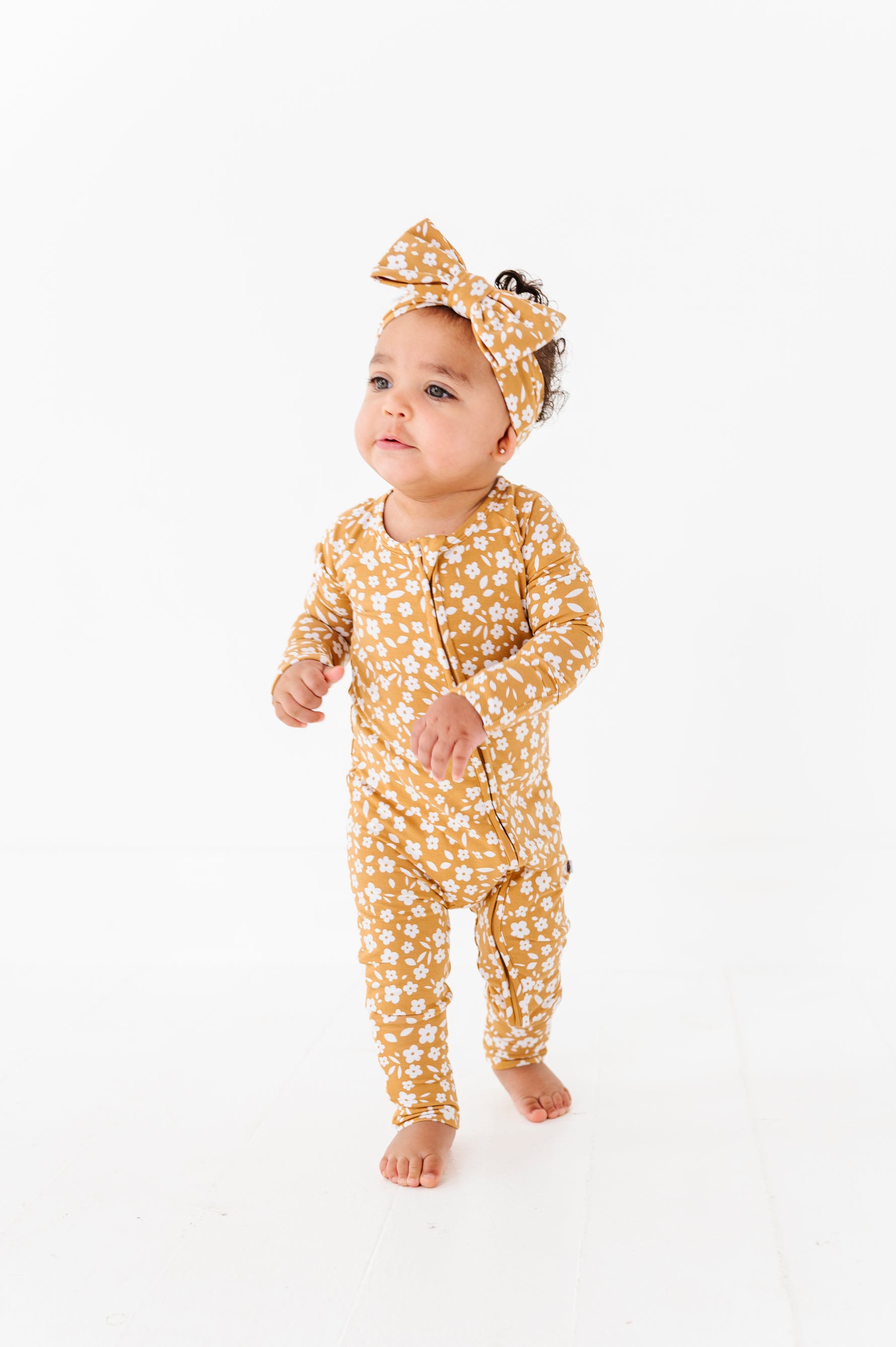 A baby in the Jammie Session Honey Blooms Romper with a matching headband stands on a white floor against a white background.
