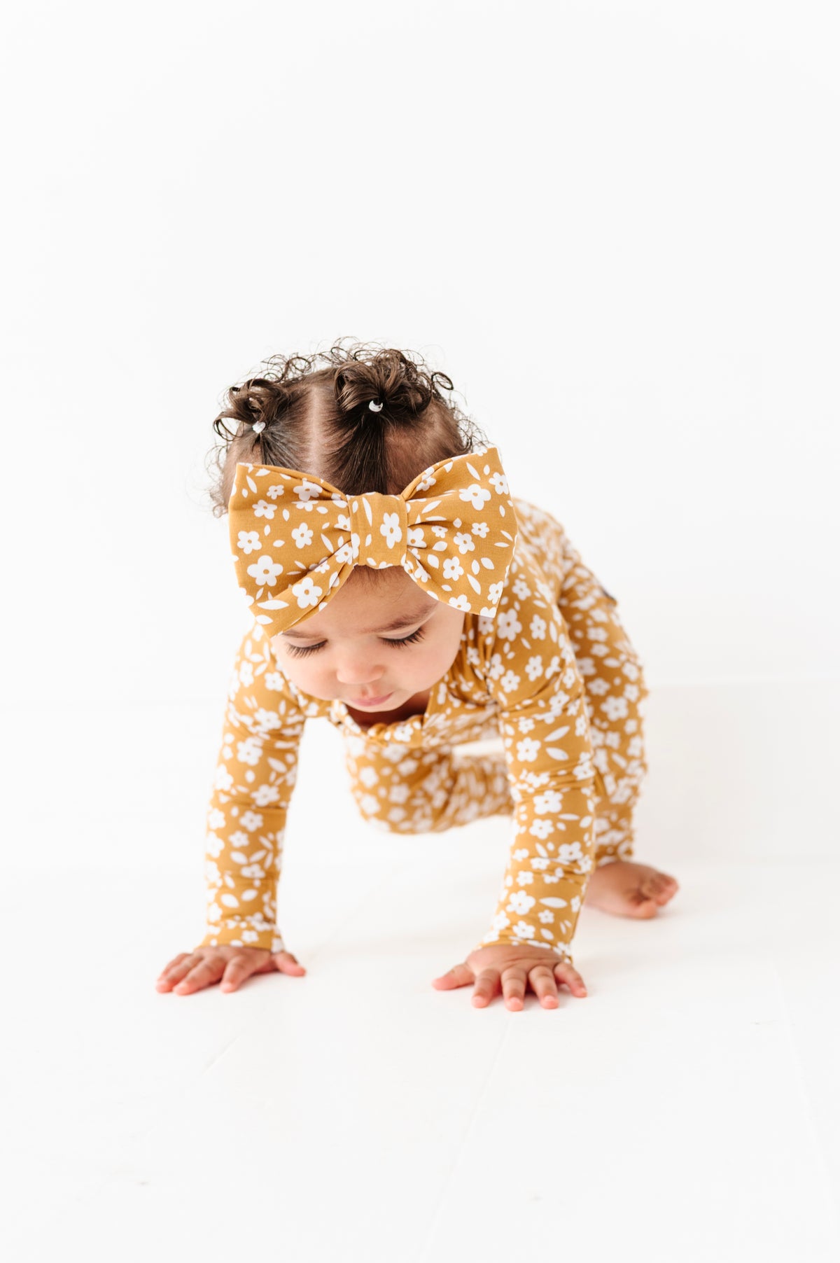 A baby wearing the Jammie Session Honey Blooms Romper in mustard yellow floral with a matching bow crawls on a white floor against a white background.