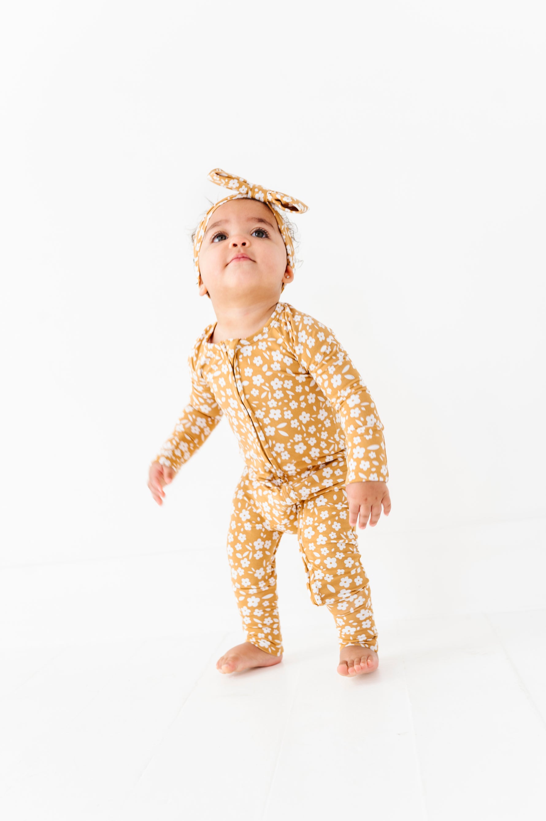 A toddler in a matching Honey Blooms Romper and headband by Jammie Session stands barefoot on a white floor, looking slightly upward.
