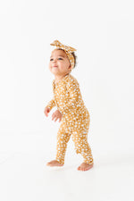 A toddler wearing the Jammie Session Honey Blooms Romper, a yellow floral bamboo outfit with a matching headband, stands barefoot on a white background.