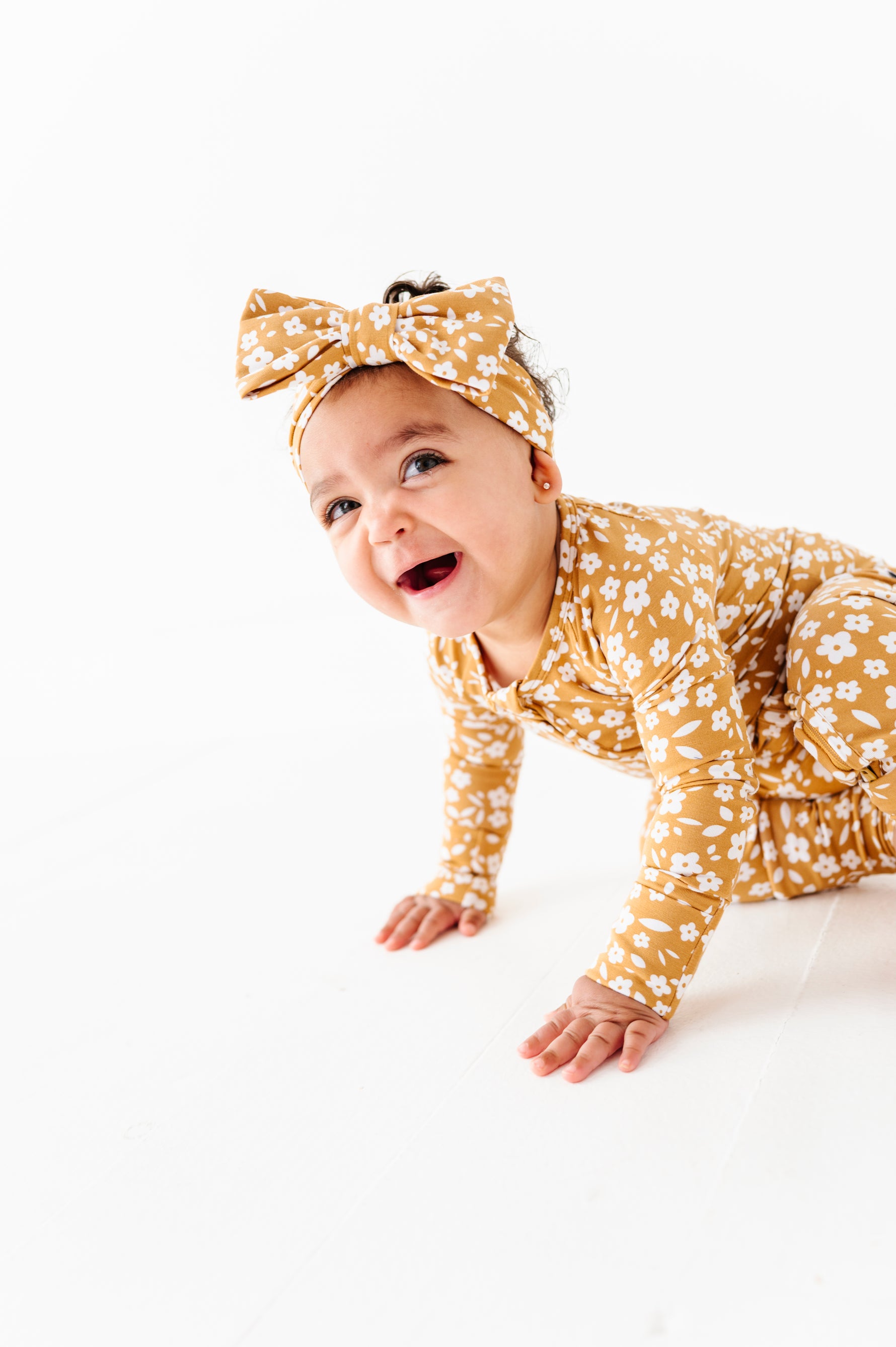 A baby in a Jammie Session Honey Blooms Romper with a matching headband crawls on a white floor, smiling and looking upward.