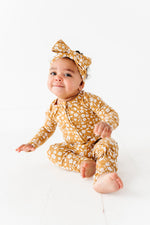 A baby sits barefoot on a white floor, looking at the camera while wearing the Jammie Session Honey Blooms Romper and a matching headband in mustard yellow floral bamboo.