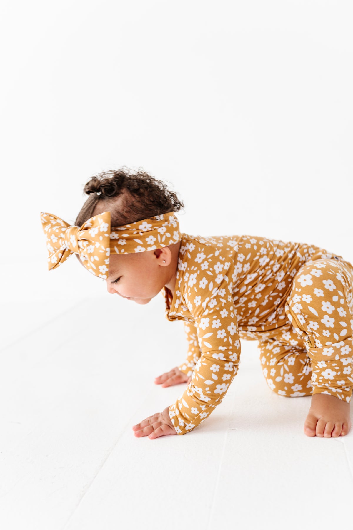 A baby in a Jammie Session Honey Blooms Romper with a matching headband crawls on a white floor against a plain white background.