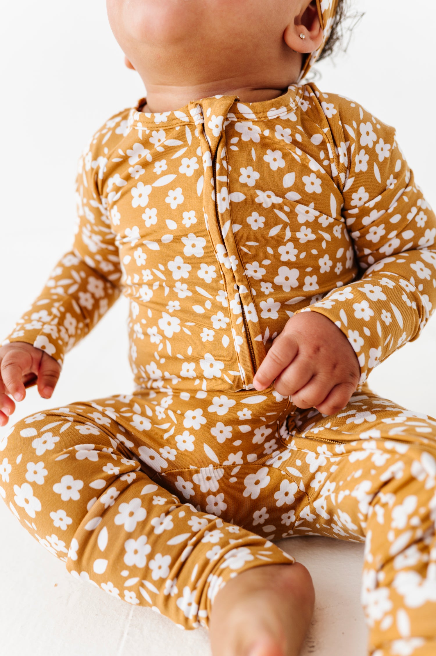 A baby sits on a white surface wearing the Jammie Session Honey Blooms Romper in mustard yellow with a floral pattern, head tilted back and face partially out of frame.