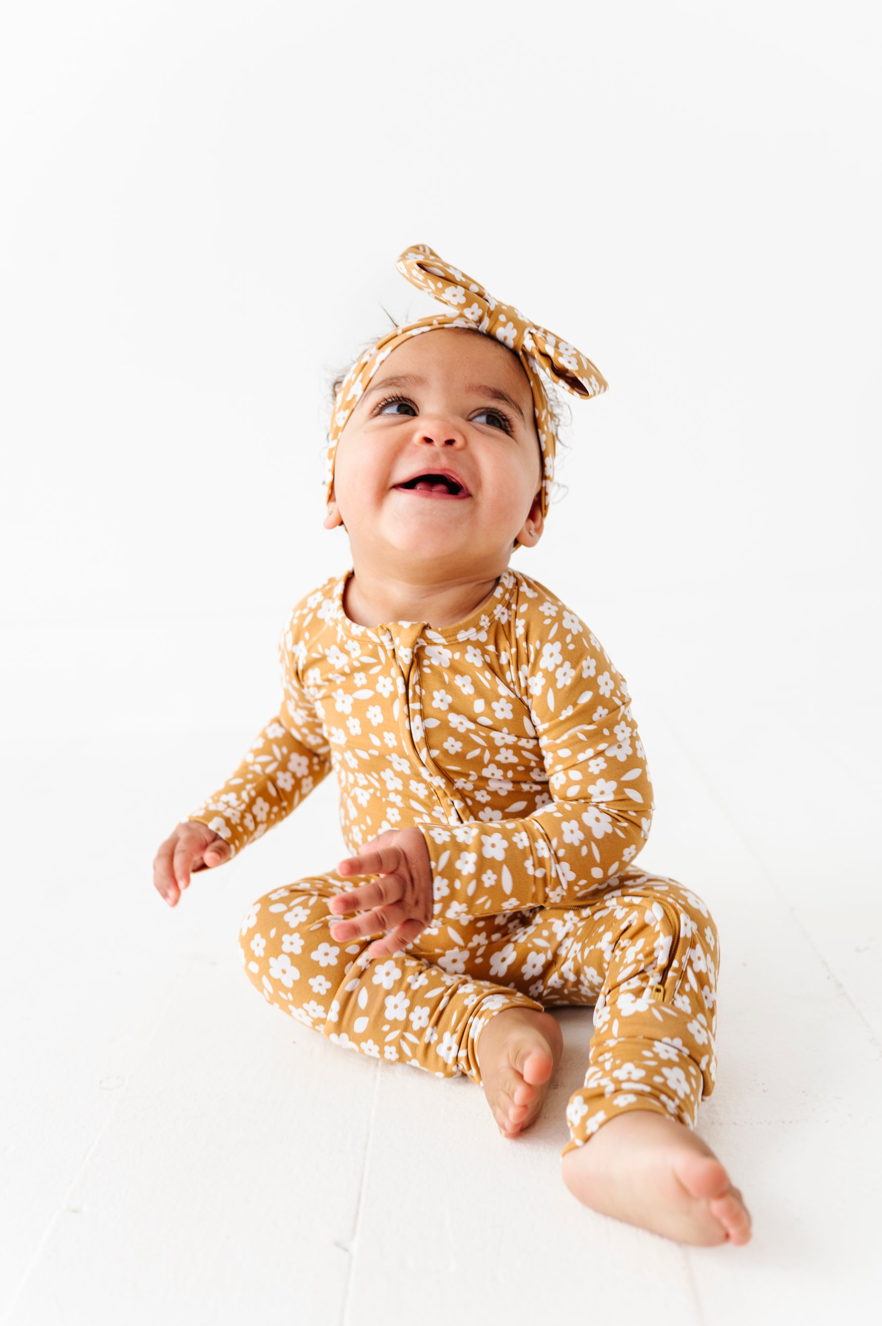 A baby sits barefoot on a white floor, smiling upward while wearing the Jammie Session Honey Blooms Romper with a matching brown floral headband.