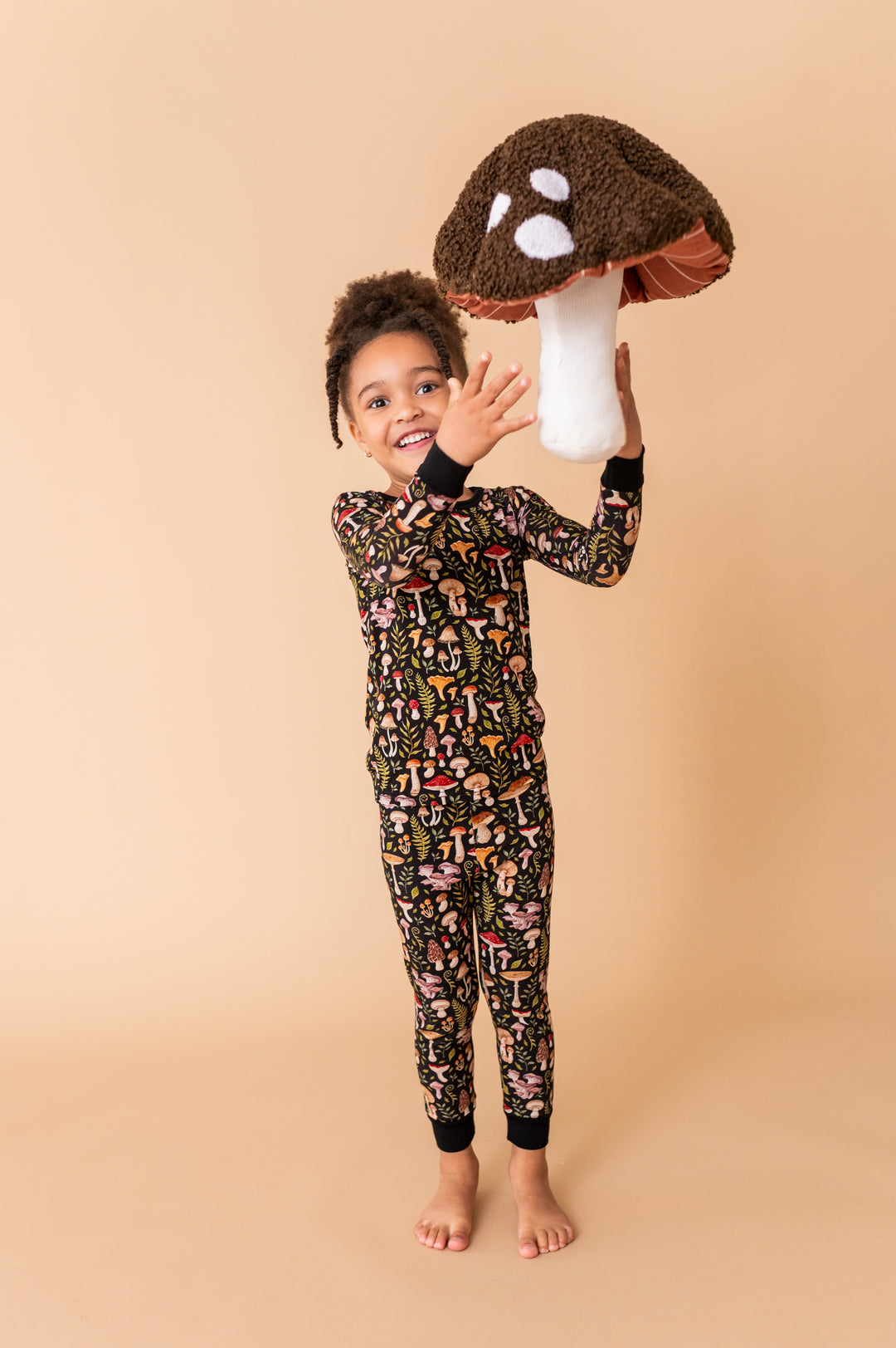 A child wearing Jammie Session's Mystic Mushrooms Long Sleeve PJ's stands barefoot, smiling and holding a large plush mushroom against a plain beige background.