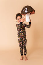 A child wearing Jammie Session's Mystic Mushrooms Long Sleeve PJ's stands barefoot, smiling and holding a large plush mushroom against a plain beige background.