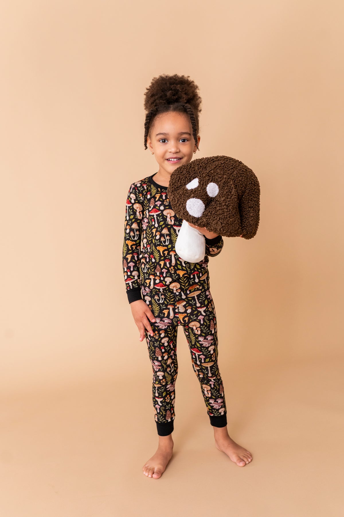 A young girl smiles at the camera, barefoot on a tan background, wearing Jammie Session's Mystic Mushrooms Long Sleeve PJ's and holding a large plush dog head.
