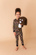 A young girl smiles at the camera, barefoot on a tan background, wearing Jammie Session's Mystic Mushrooms Long Sleeve PJ's and holding a large plush dog head.