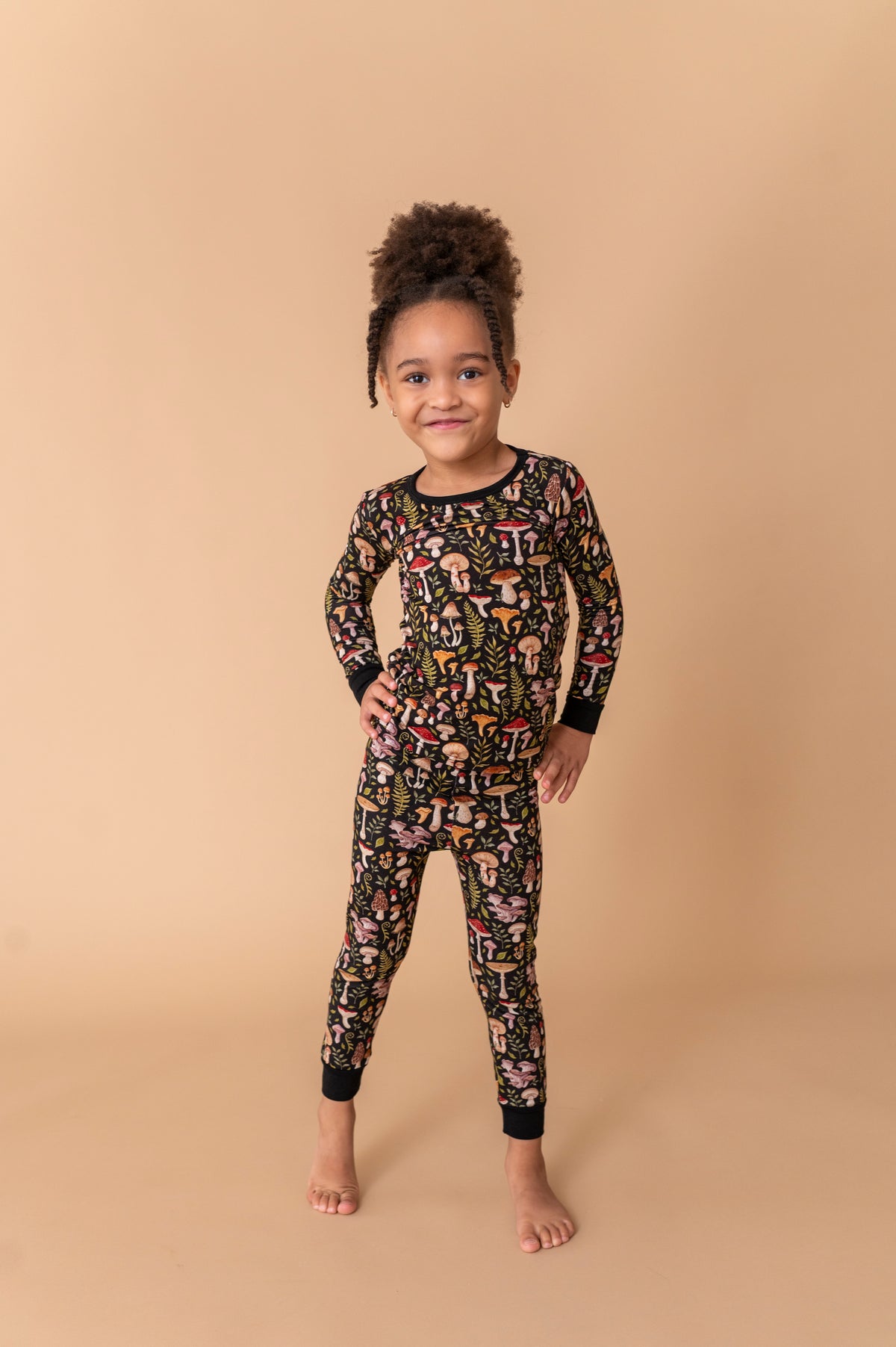 A young child stands barefoot on a tan backdrop, wearing Jammie Session’s Mystic Mushrooms Long Sleeve PJ's—a black kids’ pajama set with a colorful abstract pattern made from soft, hypoallergenic fabric.