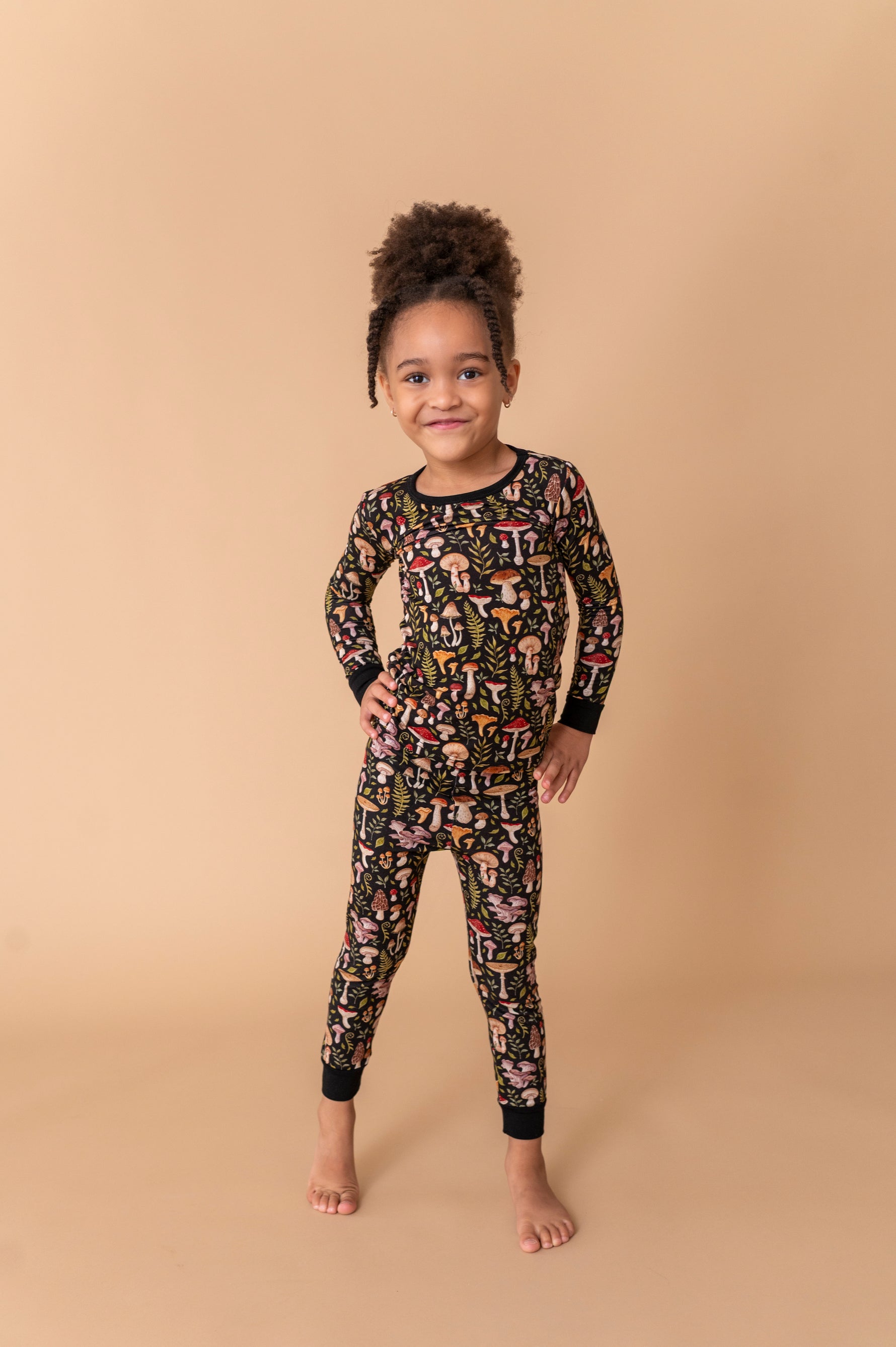 A young child stands barefoot on a tan backdrop, wearing Jammie Session’s Mystic Mushrooms Long Sleeve PJ's—a black kids’ pajama set with a colorful abstract pattern made from soft, hypoallergenic fabric.