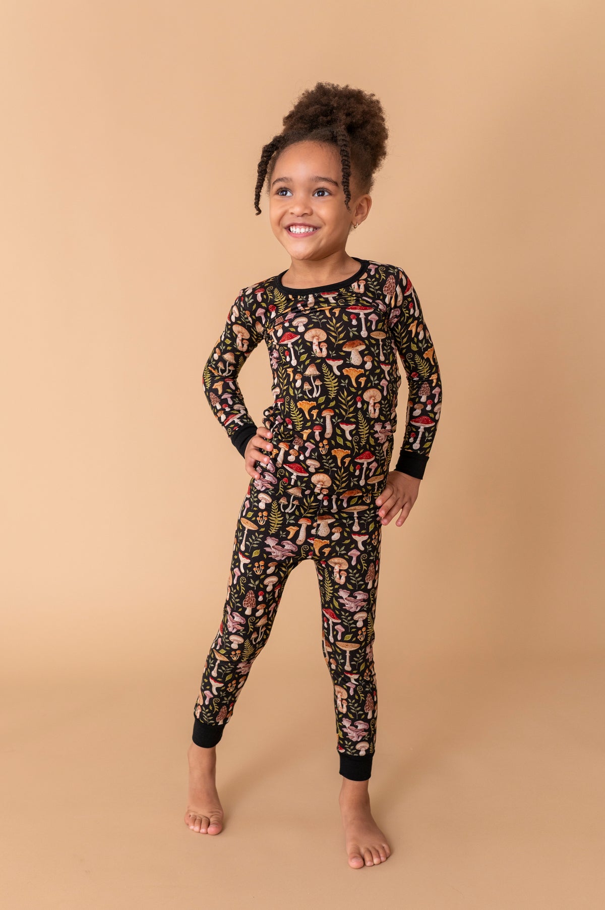 A young child stands barefoot on a tan background, wearing Jammie Session's Mystic Mushrooms Long Sleeve PJ's—colorful, cozy sleepwear perfect for kids' nights in.