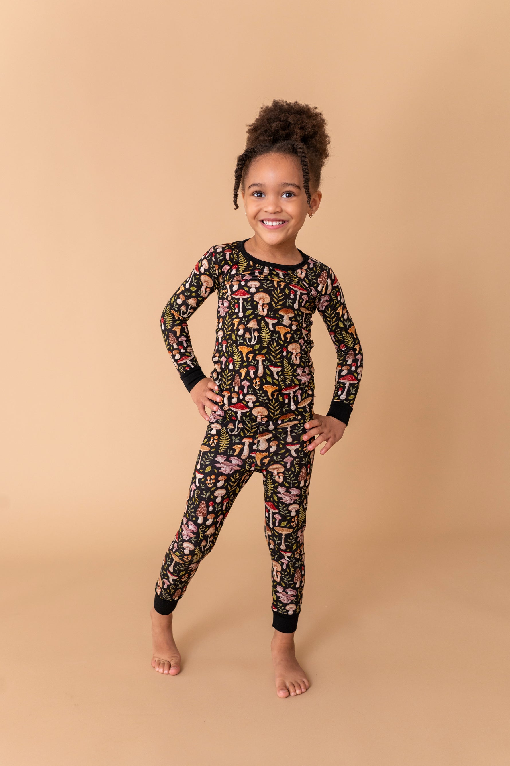 A young child smiles against a beige background, wearing Jammie Session’s Mystic Mushrooms Long Sleeve PJ’s featuring a colorful printed pattern.